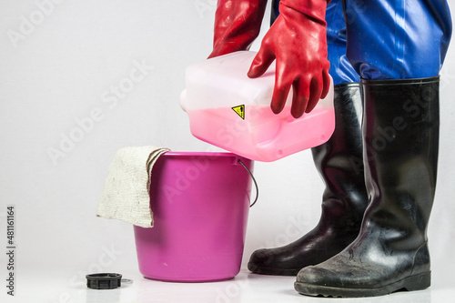 Man in a blue rubber protective suit, with  red rubber gloves and black rubber boots tilts a red, corrosive industrial cleaner in a plastic bucket.