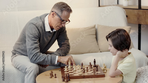 grandfather in glasses and preteen boy playing chess in living room