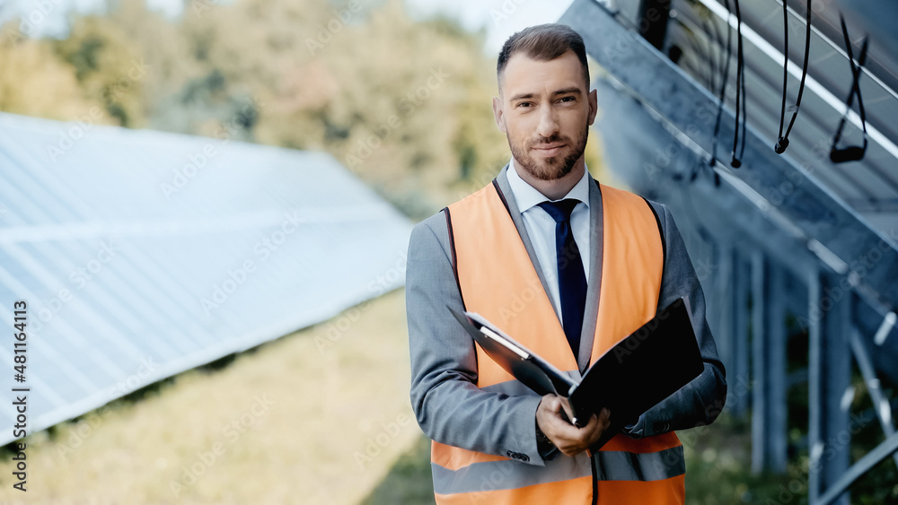 businessman in safety vest and suit holding folder near solar panels businessman in safety vest and suit holding folder near solar panels