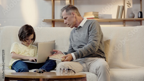 middle aged grandfather holding book near preteen grandson using laptop in living room