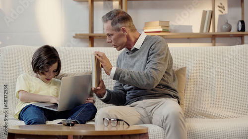 middle aged grandfather showing book to grandson using laptop in living room