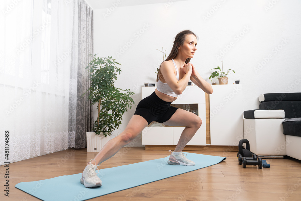 Young fitness woman in black sporty shorts doing side lunge on yoga mat during home workout in living room. Leg stretching after workout. Young fitness woman in black sporty shorts doing side lunge on yoga mat during home workout in living room. Leg stretching after workout.