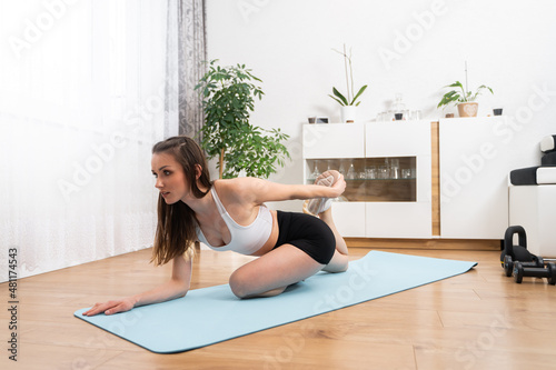 Athletic young woman in sportswear doing leg and butt stretching in living room. Attractive brunette exercising on blue mat.