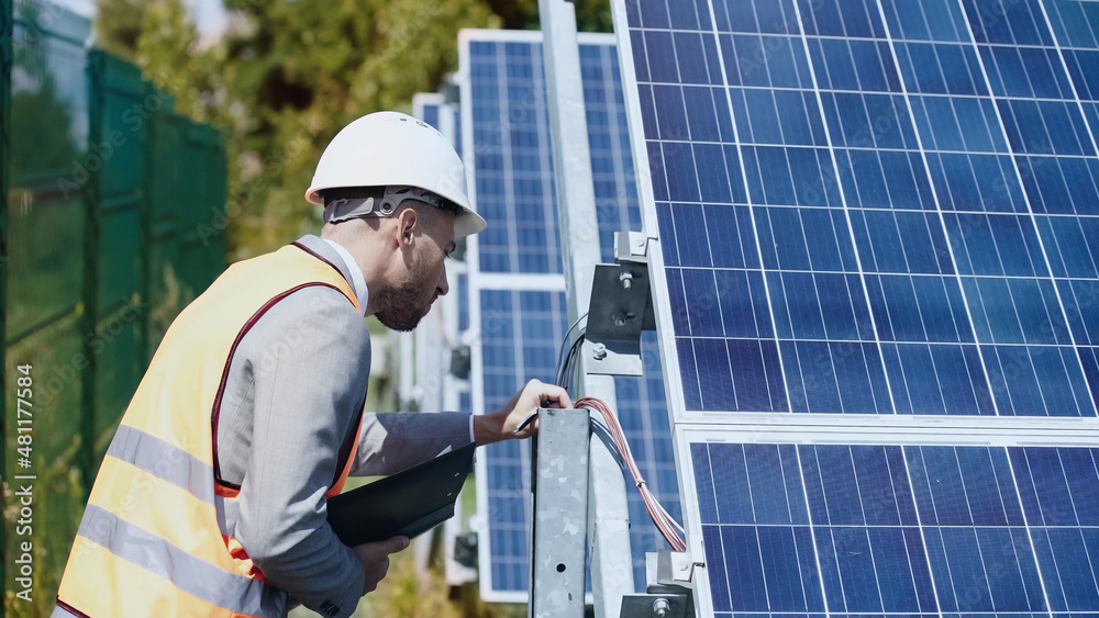 businessman in hardhat checking wires and holding folder near solar batteries outside