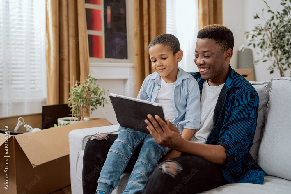 Fototapeta premium A proud father sits on the couch holding son on his lap, a tablet in hand, they take a selfie, a picture for social media, they smile