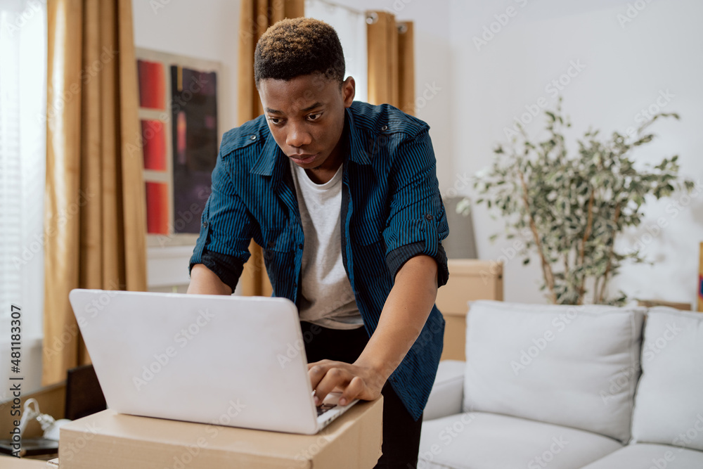 Man moves into new apartment, no furnishings except couch, laptop stands on cardboard boxes, man browses online furniture stores, looking for dresser, closet, decorations to buy Man moves into new apartment, no furnishings except couch, laptop stands on cardboard boxes, man browses online furniture stores, looking for dresser, closet, decorations to buy