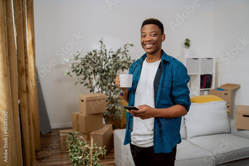 Guy is standing in the middle of the living room in a new apartment, around him are stacked boxes after the move, the man is relaxing drinking coffee in a cup and writing messages on the phone photo