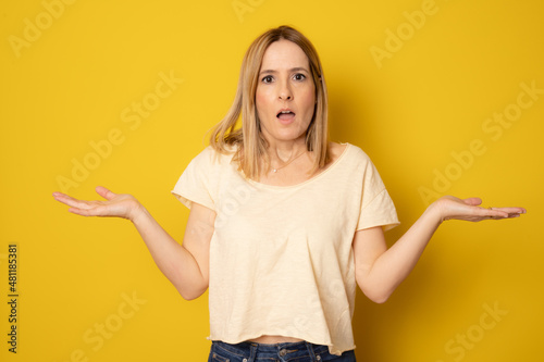 Angry woman in casual t-shirt standing with open arms isolated over yellow background.