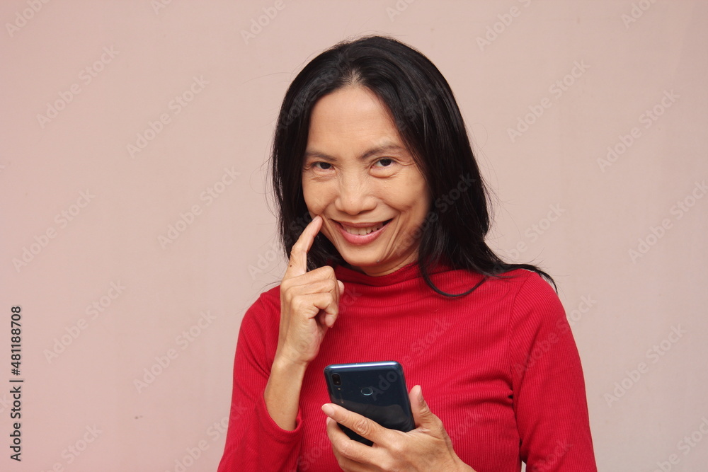 Portrait of an Asian model holding a mobile phone, turning to look at him with a smile. Wear a red sweater against a light brown background. Portrait of an Asian model holding a mobile phone, turning to look at him with a smile. Wear a red sweater against a light brown background.