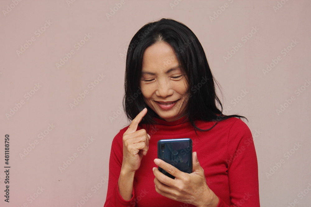 Portrait of an Asian model holding a cell phone and smiling at it. Wear a red sweater against a light brown background. Portrait of an Asian model holding a cell phone and smiling at it. Wear a red sweater against a light brown background.