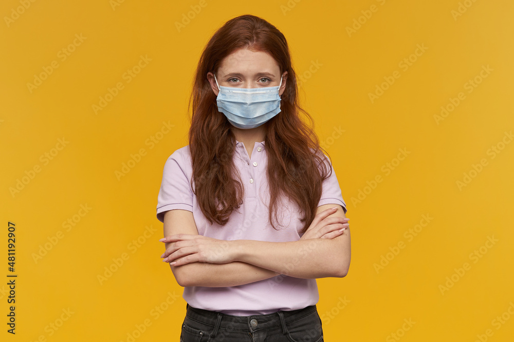 Indoor portrait of young ginger female wears medical mask looking derectly into camera with confused facial expression, keep her eyebrows raised. isolated over yellow background Indoor portrait of young ginger female wears medical mask looking derectly into camera with confused facial expression, keep her eyebrows raised. isolated over yellow background