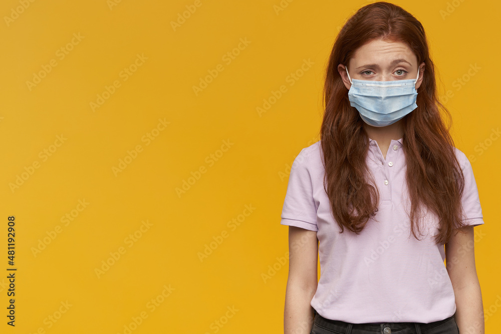 Indoor portrait of young ginger female wears medical mask looking derectly into camera with confused facial expression, keep her eyebrows raised. isolated over yellow background Indoor portrait of young ginger female wears medical mask looking derectly into camera with confused facial expression, keep her eyebrows raised. isolated over yellow background