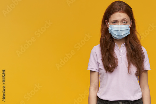Indoor portrait of young ginger female wears medical mask looking derectly into camera with confused facial expression, keep her eyebrows raised. isolated over yellow background