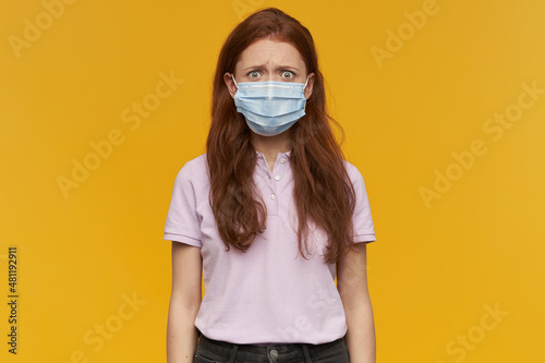 Indoor portrait of young ginger female wears medical mask looking derectly into camera with confused facial expression, keep her eyebrows raised. isolated over yellow background