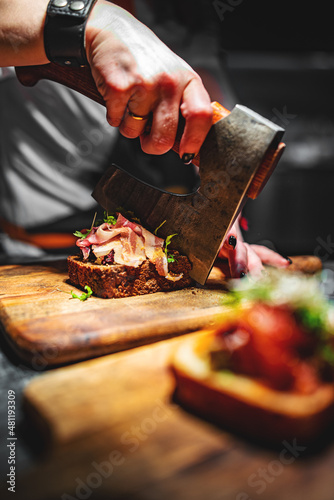 Woman chef hand cutting tasty sandwich with axe on wooden plate 