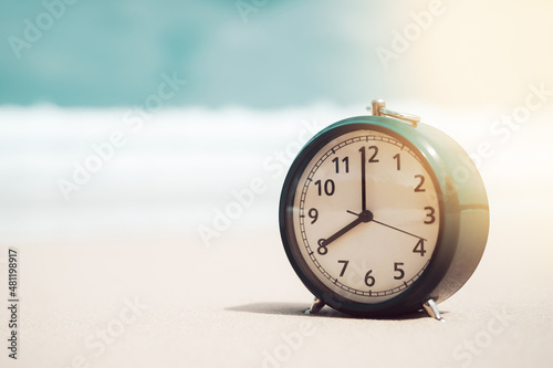 Clock on sand beach in morning suns light with smooth wave background. Copy space of time and summer holiday concept.