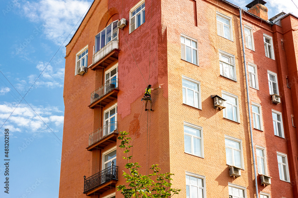Fototapeta premium A painter climber is painting the building against the blue sky with white clouds