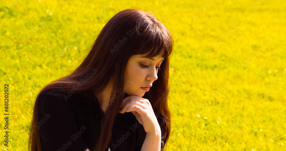 A beautiful thoughtful brunette girl sitting on the grass in the summer park A beautiful thoughtful brunette girl sitting on the grass in the summer park
