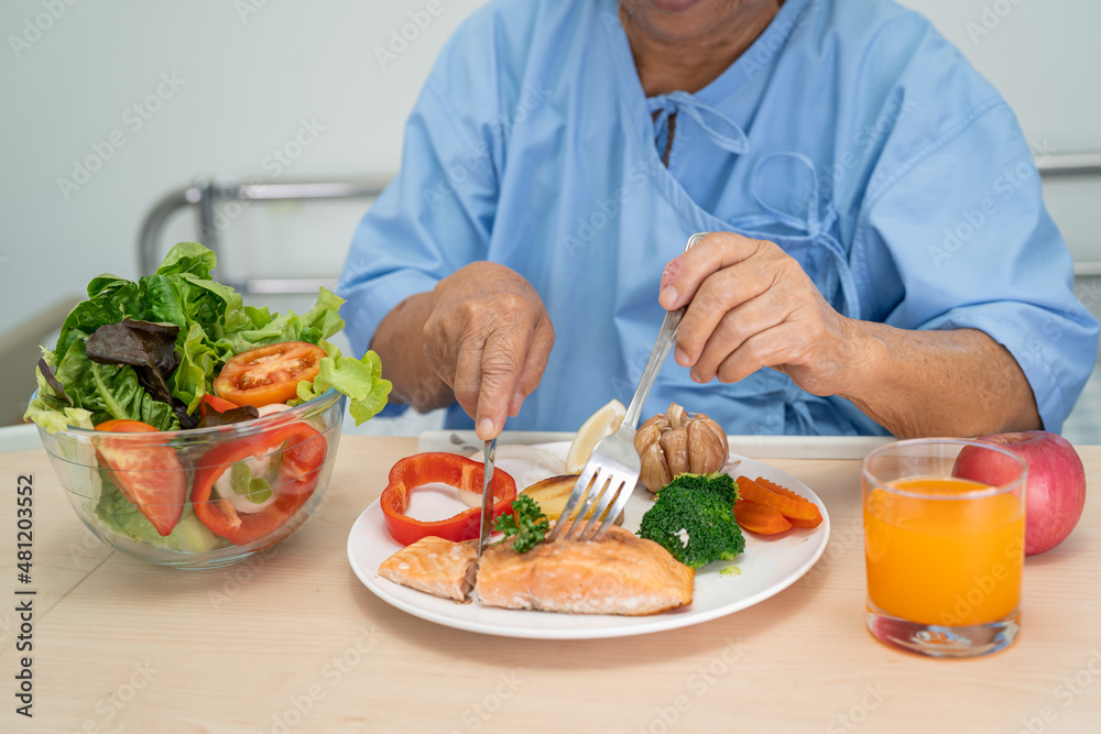 Asian senior or elderly old lady woman patient eating Salmon steak breakfast with vegetable healthy food while sitting and hungry on bed in hospital.