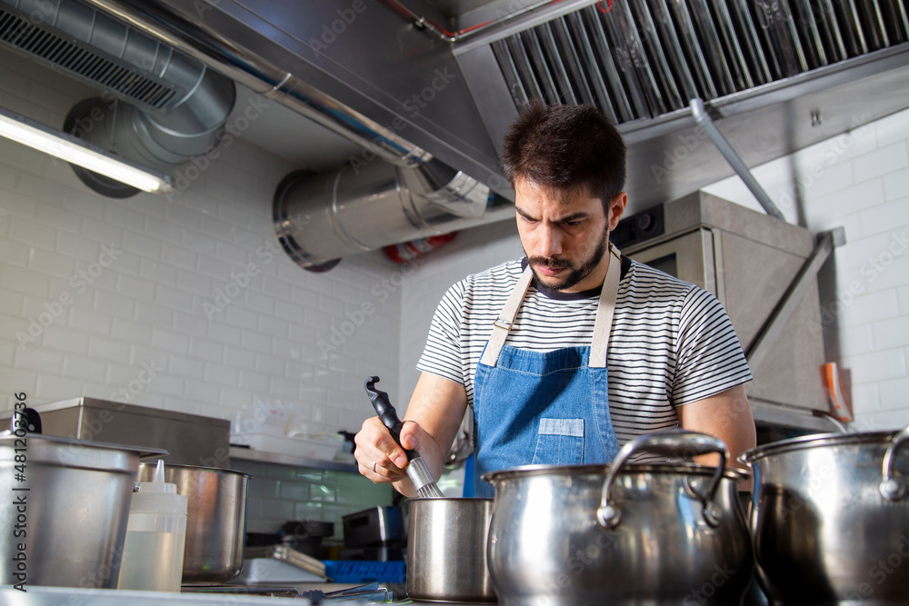Young serious ethnic male cook stirring ingredients in saucepan in kitchen Young serious ethnic male cook stirring ingredients in saucepan in kitchen
