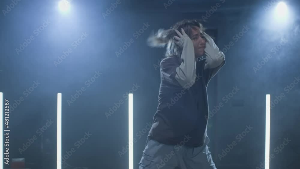 Female enjoying hip hop moves in dark studio with smoke, lighting and lightstands at background. Young woman dancing on stage. Dark studio in 4K, UHD