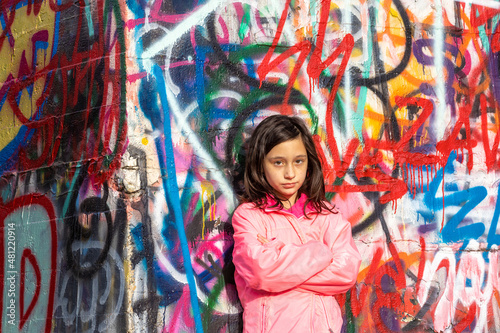 Moody pensive with deep emotion young girl with long dark hair in a pink jacket standing in front of a bright colorful graffiti wall. © Donald Blodger