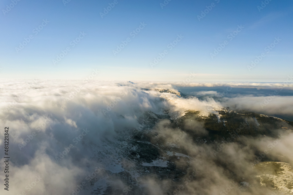 Aerial view from above of white puffy clouds covering snowy mountain tops in bright sunny day