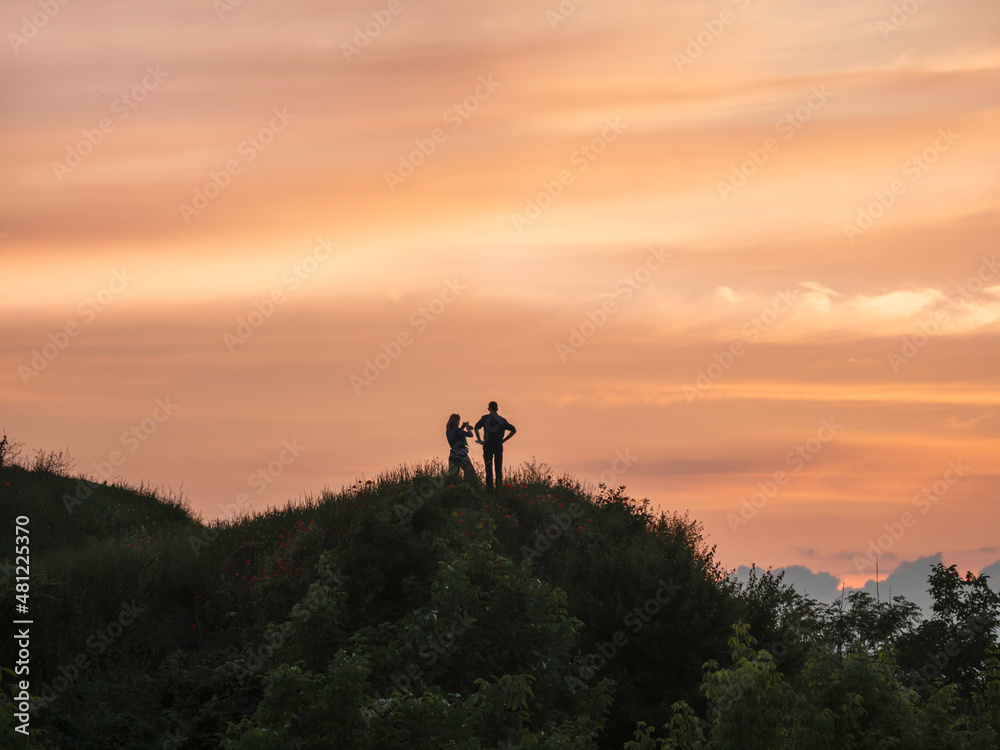 Silhouette of couple meeting sunset on hill. Woman takes picture of clouds on gorgeous orange sky background. Romantic evening outdoors.