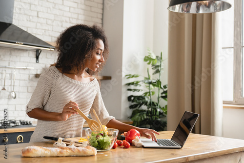 Online cooking tutorial. African young woman teenage girl using laptop for vlogging blogging looking for recipe while cooking vegetable vegan salad meal, healthy eating habits.