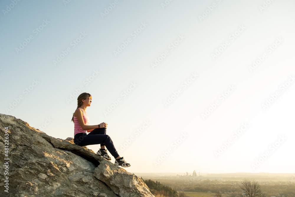 Woman hiker sitting on a steep big rock enjoying warm summer day. Young female climber resting during sports activity in nature. Active recreation in nature concept.
