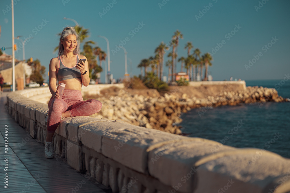 Active woman in sports clothes sitting by the seashore, holding mobile phone and lookind at it