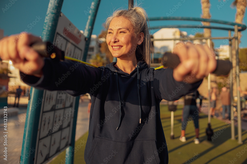 Obraz premium Portrait of sporty woman training with rubber bands at outdoor sportsground, smiling looking to the side