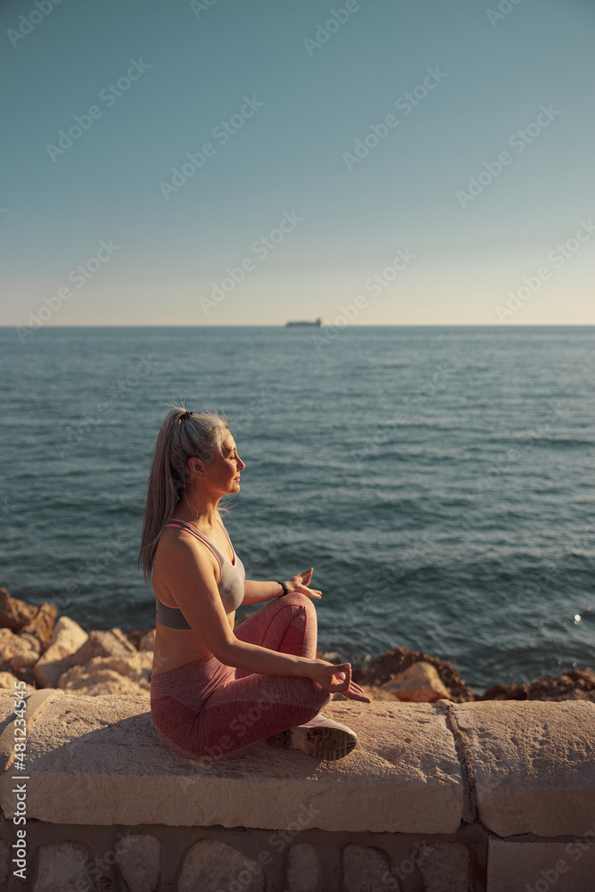 Female in sports clothes sitting by the beautiful seashore in lotus position and training on sunny day