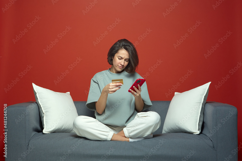 Upset frowning young girl internet shopper sitting on couch hold phone, credit card suffer of overspending money