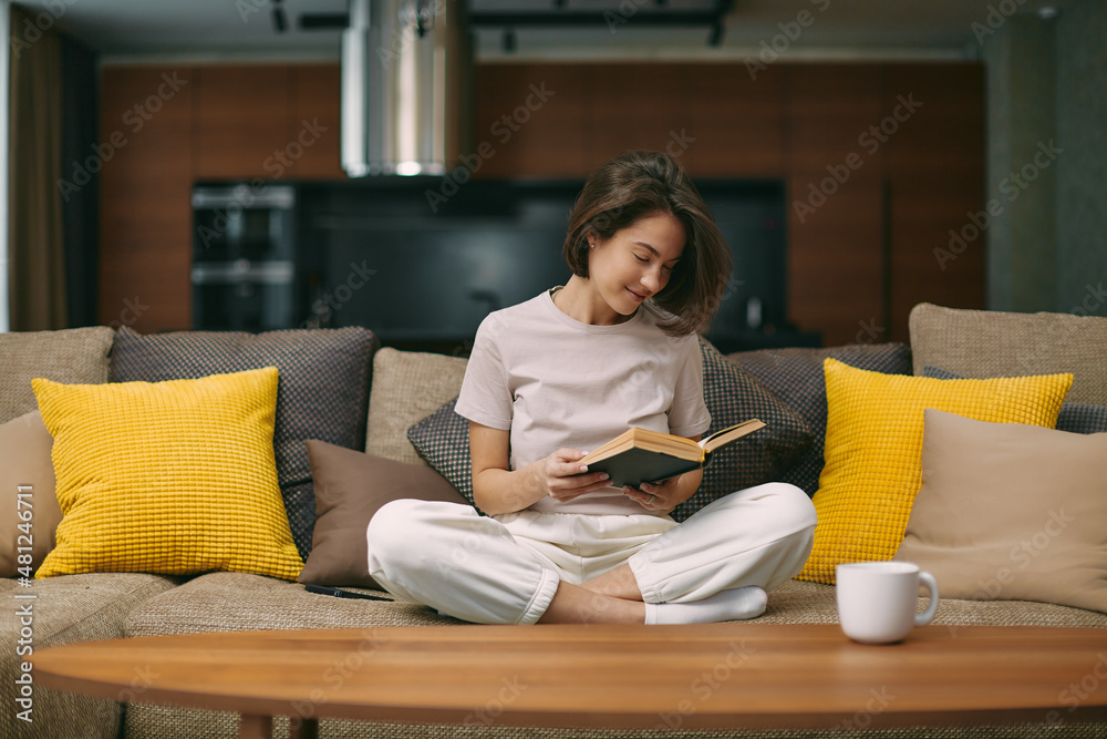 Smiling young girl student reading paper book rest sitting on sofa in living room at home enjoy novel. Self education Smiling young girl student reading paper book rest sitting on sofa in living room at home enjoy novel. Self education