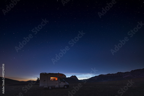 Panoramic night sky over Campo Imperatore highlands, Abruzzo, Italy. The Milky Way galaxy arc and stars over illuminated camper van. Camping freedom in unique hills landscape.