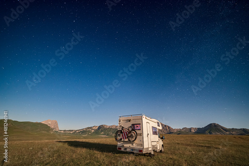 Panoramic night sky over Campo Imperatore highlands, Abruzzo, Italy. The Milky Way galaxy arc and stars over illuminated camper van. Camping freedom in unique hills landscape. photo