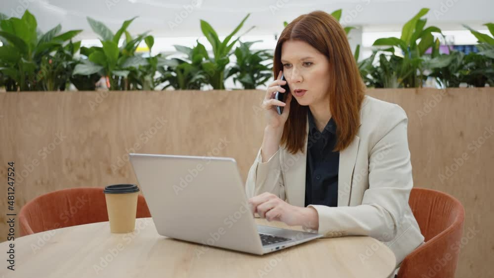Businesswoman talking on phone and using laptop in open work space, carton coffee cup standing beside, green plants on wall behind her, remote worker in cafe, freelancer Businesswoman talking on phone and using laptop in open work space, carton coffee cup standing beside, green plants on wall behind her, remote worker in cafe, freelancer