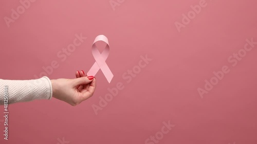 Closeup of woman hand holding pink ribbon, symbol of breast cancer awareness, oncological disease prevention and female healthcare. Indoor studio shot isolated on pink background.