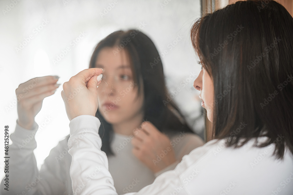 A young woman stands in front of a mirror and looks in surprise at the lost hair in her hands, the problem of hair loss after covid disease A young woman stands in front of a mirror and looks in surprise at the lost hair in her hands, the problem of hair loss after covid disease