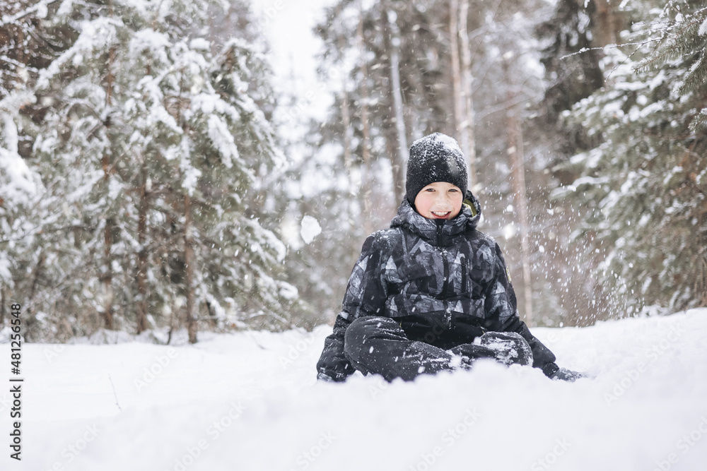 Happy teenager boy sitting on snow in winter forest. Child having fun outdoors. Joyful adolescent playing in snow at snowfall. Laughing smiling kid walking in winter park in cold weather Happy teenager boy sitting on snow in winter forest. Child having fun outdoors. Joyful adolescent playing in snow at snowfall. Laughing smiling kid walking in winter park in cold weather