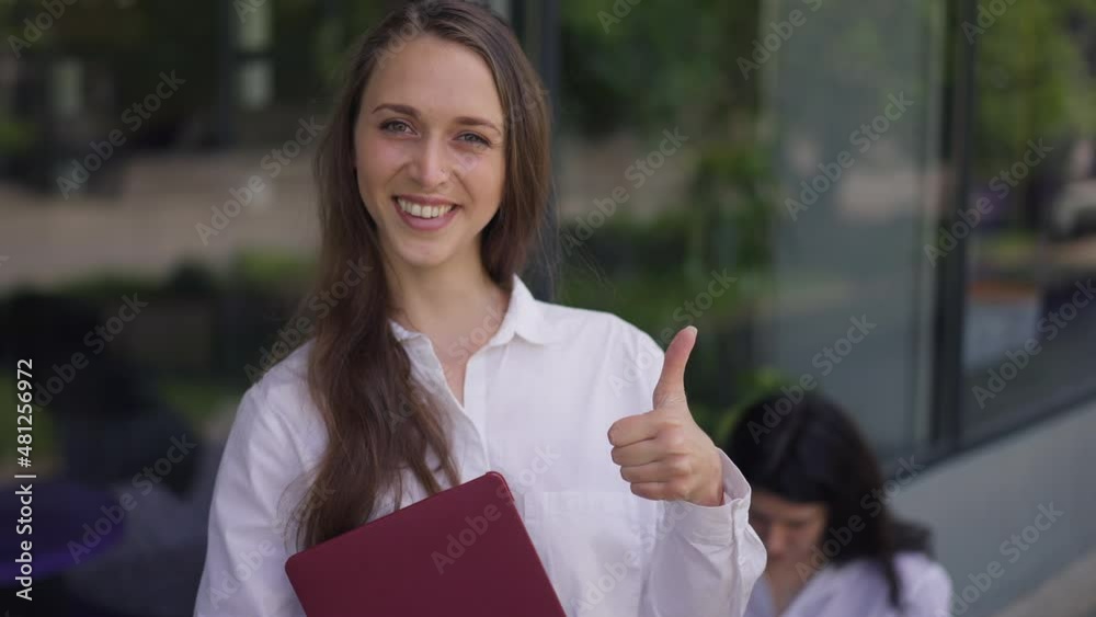 Positive woman with laptop gesturing thumb up smiling looking at camera standing on city street with blurred friend sitting at background. Portrait of confident happy millennial lady posing outdoors Positive woman with laptop gesturing thumb up smiling looking at camera standing on city street with blurred friend sitting at background. Portrait of confident happy millennial lady posing outdoors