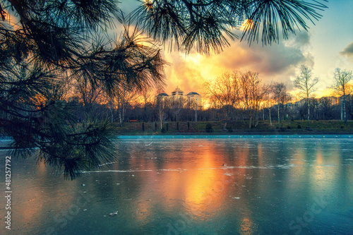 Winter city park at sunset and a frozen lake with trees without leaves