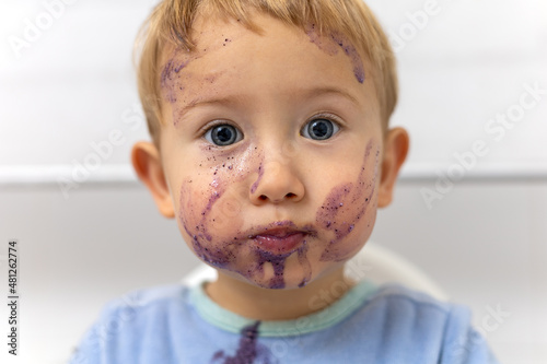 Primer plano de un niño rubio de ojos azules desayunando con la cara sucia de arandanos.