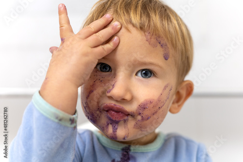 Primer plano de un niño de 2 años rubio con ojos azules se ensucia la cara desayunando