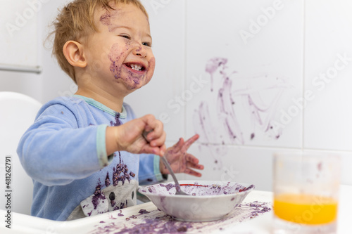 Niño rubio con pijama azul se rie después de manchar toda la mesa con su desayuno