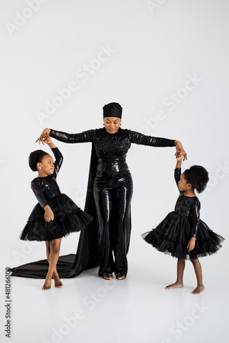 African woman in elegant black outfit and hat holding hands with her graceful little daughters in stylish dresses over white background. Proud of being mother of two female kids.