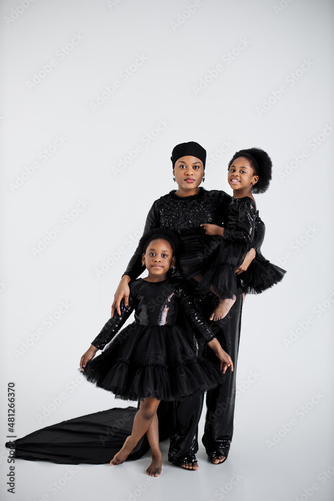 Two cute african sisters standing in embrace with their charming mother over white studio background. Stunning woman with daughters wearing elegant evening clothes.