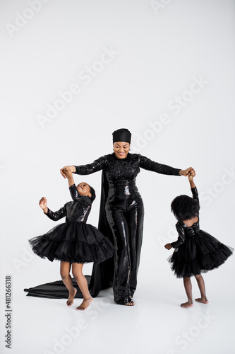 African islamic woman in elegant black outfit and hat holding hands with her graceful little daughters in stylish dresses over white background. Proud of being mother of two female kids. © sofiko14