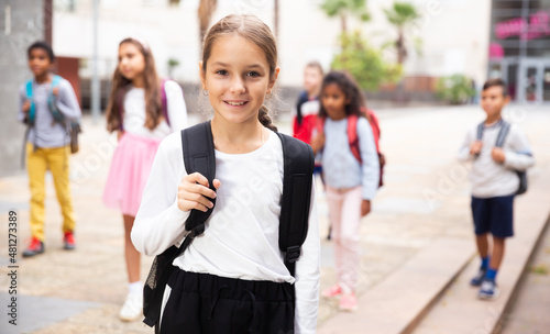 Portrait of positive schoolgirl standing near school, children on background